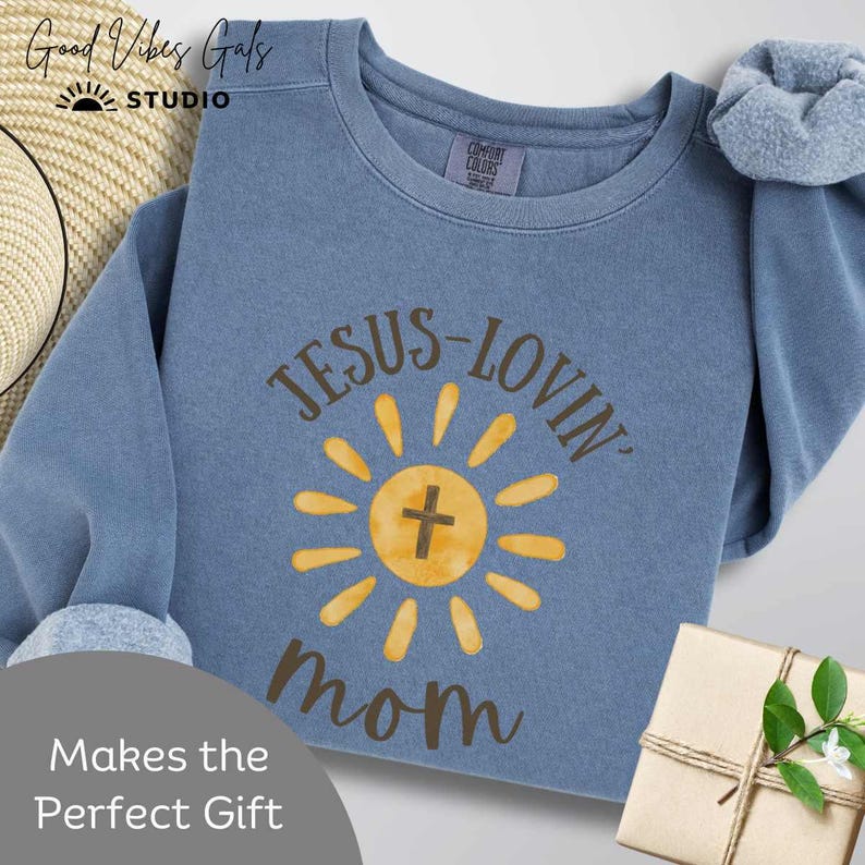 May include: A blue crew neck sweatshirt with the text "JESUS-LOVIN' mom" in brown lettering. The design includes a yellow sun with a cross in the center. The brand "Good Vibes Gals STUDIO" is at the top.