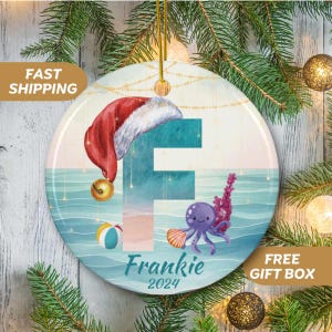 May include: A round, white ceramic ornament with a blue and green ocean scene. The letter F is in the center of the ornament, with a purple octopus and a red Santa hat. The text "Frankie 2024" is at the bottom of the ornament.