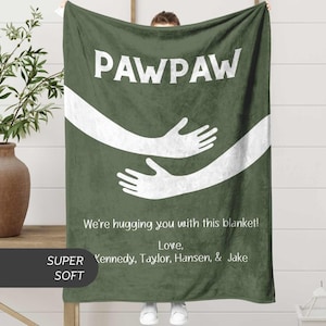 May include: A dark green blanket with the word "PAWPAW" in white. The blanket features a white graphic of two embracing arms. Text below reads "We're hugging you with this blanket! Love, Kennedy, Taylor, Hansen, & Jake."