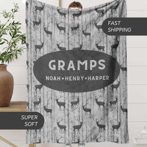 May include: A gray and white blanket featuring a forest scene with deer silhouettes. The blanket has an oval design with the word "GRAMPS" and the names "NOAH, HENRY, HARPER". The blanket is labeled "SUPER SOFT".