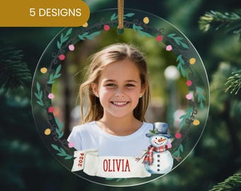 May include: A round, clear, acrylic Christmas ornament with a green wreath design, a snowman, and a photo frame. The ornament has a white banner with the text "2024" and "OLIVIA" in red.