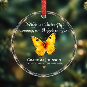 May include: A round, clear glass ornament with a faceted edge, featuring a yellow butterfly graphic and the text "When a Butterfly appears an Angel is near." Below, it reads "GRANDMA JOHNSON" and "JULY 12TH, 1932 - JUNE 11TH, 2026" with a small gold heart.