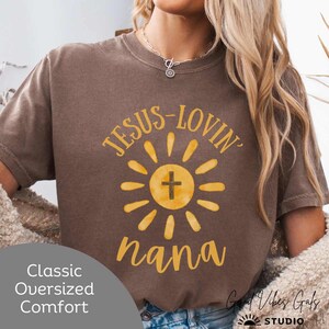 May include: Brown t-shirt with the text "JESUS-LOVIN' nana" in yellow. The design features a yellow sun with a cross in the center. The shirt has short sleeves and a crew neck. The words "Classic Oversized Comfort" are printed on the bottom left.