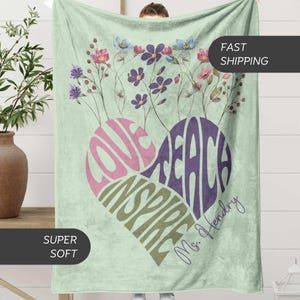 May include: A soft, light green blanket with a heart-shaped design. The heart is filled with the words "LOVE, TEACH, INSPIRE" in pink, purple, and green. Floral accents and the name "Ms. Hendry" are also featured.