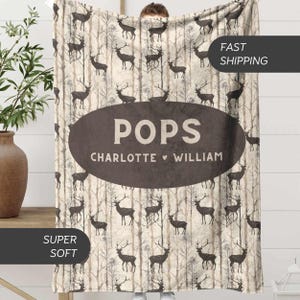 May include: A beige blanket with a repeating pattern of deer and trees. The blanket has a large brown oval with the word "POPS" and the names "Charlotte" and "William". The blanket is labeled "SUPER SOFT".