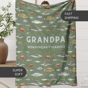 May include: A green blanket with a fish pattern and the word "GRANDPA" in white. The names "NOAH", "HENRY", and "HARPER" are below the word "GRANDPA". The blanket has a soft texture.