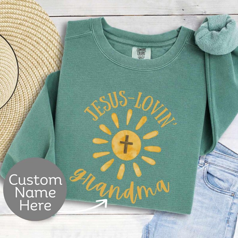 May include: A teal-colored sweatshirt with the words "JESUS-LOVIN' grandma" in yellow script. The design includes a yellow sunburst with a cross in the center. The sweatshirt is folded, with a portion of a straw hat and jeans visible.