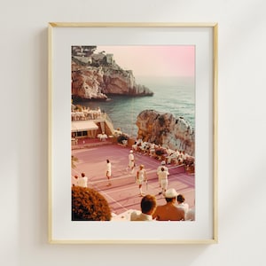 May include: Framed print of a vintage tennis match on a pink court by the sea. Players in white attire are visible. The background features a rocky coastline, turquoise water, and a pink sky. The print is in a light wood frame.