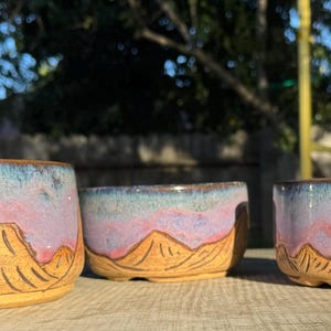 May include: Three ceramic bowls with a mountain design. The bowls have a light brown base, a pink and purple middle, and a blue top. The mountain design is etched into the bowls. The bowls are of varying sizes.