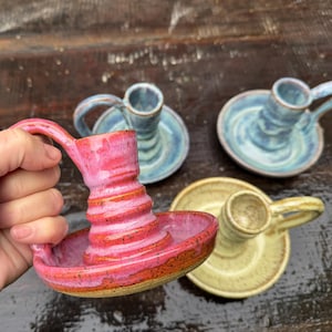 May include: Handmade ceramic candle holders in various colors. One is held in a hand, showing a vibrant pink with orange accents. Other candle holders are blue and yellow, each with a handle.