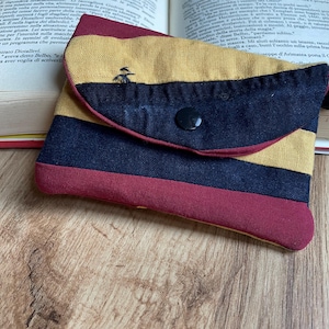Può includere: Piccola pochette a righe con chiusura a patta. La pochette presenta strisce orizzontali blu navy, giallo senape e bordeaux. Un piccolo bottone nero chiude la patta. È visibile un piccolo disegno ricamato.