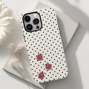 May include: A white iPhone case with a black polka dot pattern and three red ladybug illustrations. The case has a black camera lens border and the text "AdmireYourDreams" in the bottom right corner.