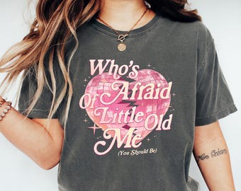May include: Dark gray t-shirt with the text "Who's Afraid of Little Old Me" in pink, set within a heart-shaped disco ball design. The shirt is paired with blue jeans and white sneakers.