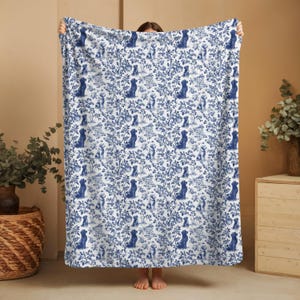 May include: A white blanket with a blue toile pattern featuring dogs, trees, and floral elements. The blanket is held up, showcasing the full design. The overall theme is a decorative home textile.