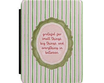 May include: A rectangular e-reader case featuring a vertical pink and green striped pattern. Centered is an ornate olive green frame surrounding a light pink oval containing the red text: grateful for small things. big things. and everything in between.