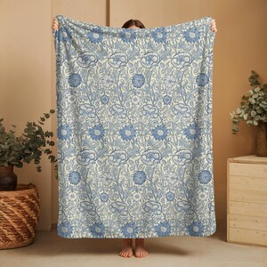 May include: A large, soft blanket with a repeating floral pattern in shades of light and medium blue on a cream background. The design features stylized flowers and foliage, creating a classic, elegant look. The blanket is held up, showcasing its full size.