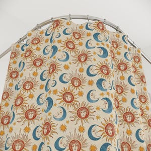 May include: Cream-coloured shower curtain featuring a repeating pattern of suns, moons, and stars. The suns have faces, and the moons are crescent-shaped. The colours are orange, blue, and yellow. The curtain hangs from a curved metal rail.