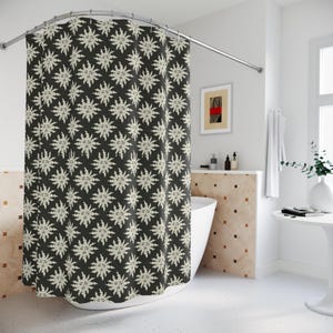 May include: A dark grey shower curtain featuring a repeating pattern of off-white sun-like faces. The curtain hangs in a bathroom with a white bath and a small table with a vase.