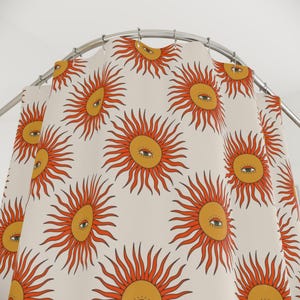May include: Cream shower curtain with a repeating pattern of stylized suns. Each sun has a yellow centre with an eye and orange rays. The curtain is hanging from a silver curved shower rail.