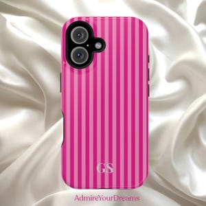 May include: A pink phone case with vertical stripes in varying shades of pink. The case has a camera cutout and black side buttons. The initials "GS" are in silver at the bottom. The text "AdmireYourDreams" is at the bottom.