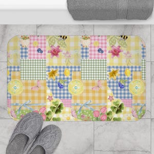 May include: A rectangular bath mat featuring a patchwork design of yellow, blue, and green gingham squares. The pattern includes illustrations of pink lilies, yellow lemons, blue berries, green clover leaves, butterflies, and light blue ribbons.