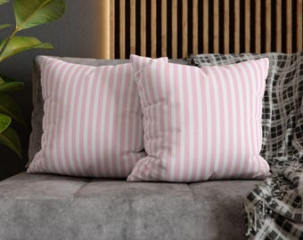 May include: Two square throw pillows with vertical pink and white stripes rest on a gray sofa. A patterned blanket is draped on the sofa. The pillows add a touch of color and pattern to the neutral-toned living space.