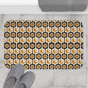 Könnte beinhalten: Rechteckige Badematte mit einem sich wiederholenden Muster aus stilisierten Sonnen, Monden und Sternen in Orange und Creme auf schwarzem Hintergrund. Die Matte liegt auf einem gefliesten Boden, graue Hausschuhe sind sichtbar.