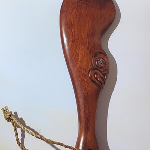 May include: A wooden carving of a face with a brown rope attached to the bottom. The carving is in the shape of a paddle.