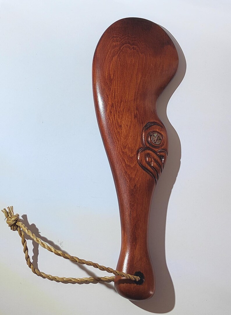 May include: A wooden carving of a face with a brown rope attached to the bottom. The carving is in the shape of a paddle.