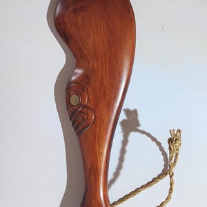 May include: A wooden carving of a Maori design with a brown stain finish. The carving has a rope loop attached to the bottom.