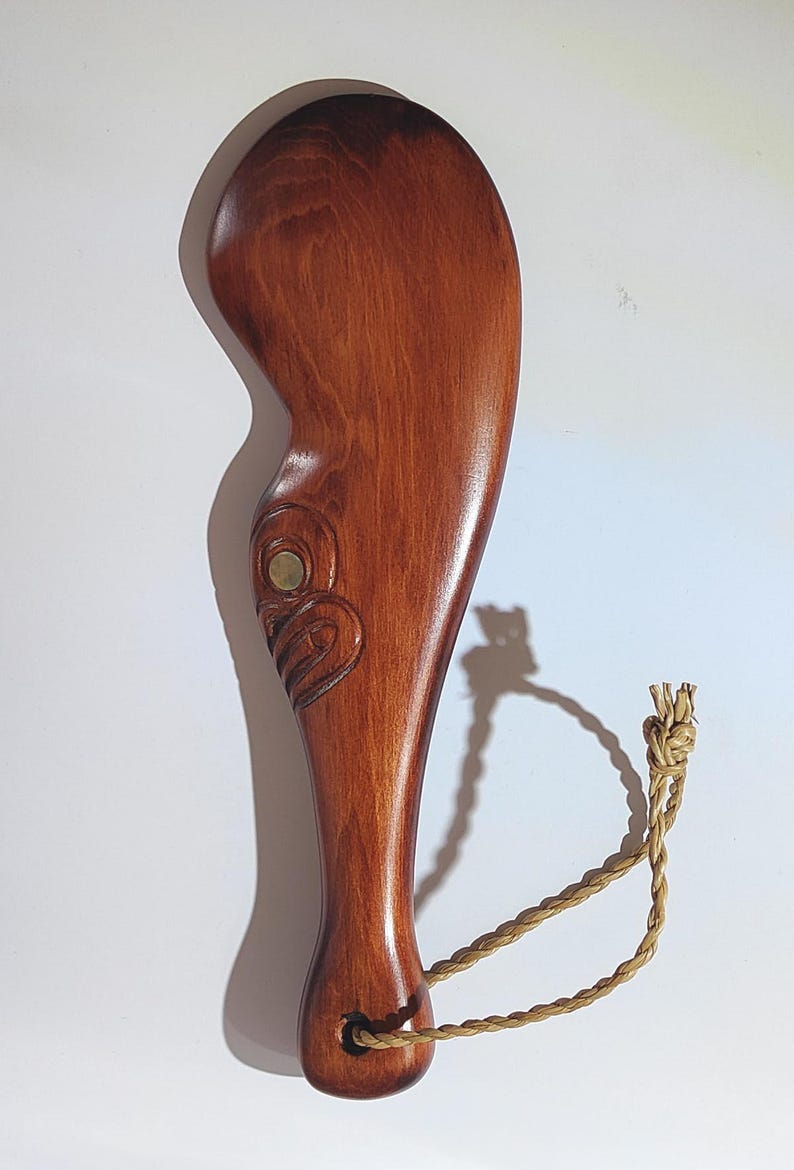 May include: A wooden carving of a Maori design with a brown stain finish. The carving has a rope loop attached to the bottom.