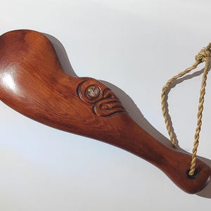 May include: A wooden carving of a Maori design with a rope handle. The carving is brown and has a white shell inlay.