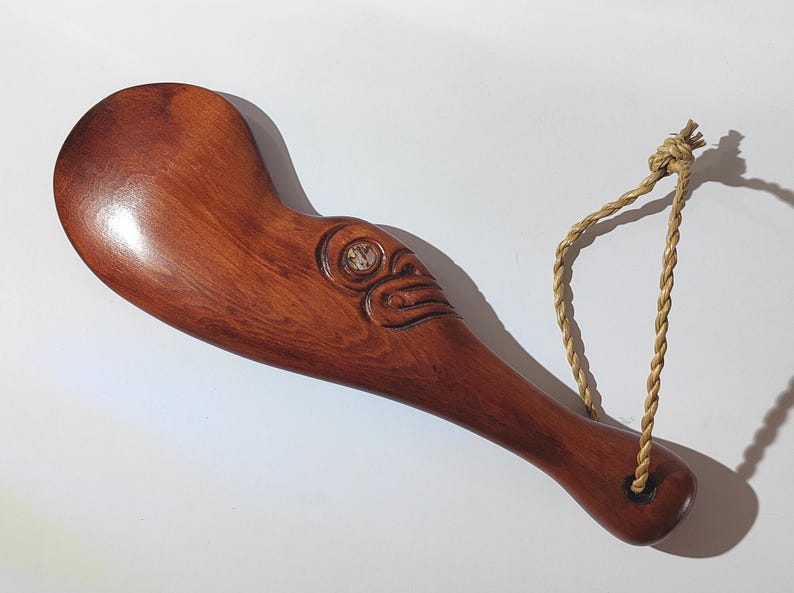 May include: A wooden carving of a Maori design with a rope handle. The carving is brown and has a white shell inlay.