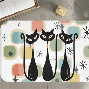 Può includere: Un tappetino da bagno bianco con un design retrò che presenta tre gatti neri con code lunghe e forme geometriche colorate. Lo sfondo è bianco con un motivo a stelle.