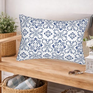 Pode incluir: Uma almofada retangular com fundo branco e um padrão repetitivo de flores e volutas em azul escuro. A almofada está posicionada horizontalmente, exibindo um design simétrico de pétalas estilizadas e vinhas intrincadas.