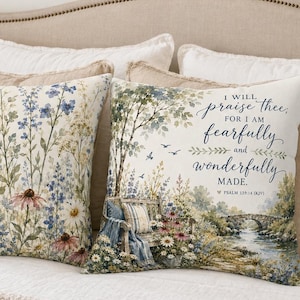 May include: Two square throw pillows featuring watercolour-style illustrations. One pillow displays a field of wildflowers in blue, pink, and yellow. The other pillow depicts a garden scene with a bench, a stream, and the text I WILL PRAISE THEE, FOR I AM FEARFULLY AND WONDERFULLY MADE. PSALM 139:14 (KJV).