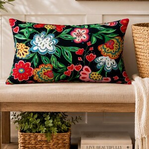 May include: A rectangular throw pillow featuring a vibrant floral pattern with red, blue, yellow, and pink flowers and green leaves set against a solid black background. The pillow is positioned on a light beige upholstered bench seat.