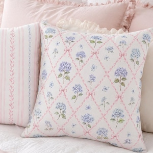 Könnte beinhalten: Zwei quadratische Dekokissen auf einem Bett. Ein Kissen hat einen weißen Hintergrund mit vertikalen rosa Streifen und blauen Blumenranken. Das andere Kissen zeigt einen weißen Hintergrund mit einem rosa Rautenmuster und blauen Hortensien.