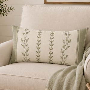 May include: A rectangular lumbar cushion featuring a cream-coloured fabric base with vertical rows of embroidered green leaf vines. The cushion has solid green fabric borders on the left and right sides, creating a structured and symmetrical botanical design.