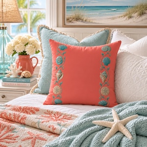 May include: A square coral-colored throw pillow featuring two vertical embroidered bands of teal and beige seashells and sand dollars. The pillow has a textured fabric finish and is positioned among other pillows on a bed with a white quilt.