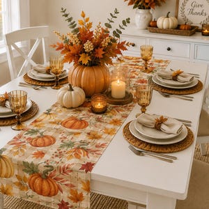 Op de afbeelding: Een gedekte eettafel met een tafelloper met pompoen- en herfstbladpatroon, witte borden, amberkleurige glazen, gevlochten placemats en een middelpunt van een grote pompoen omringd door herfstbladeren en kaarsen.