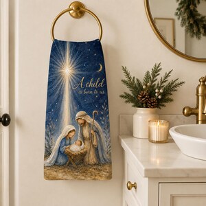 May include: A rectangular hand towel hanging on a gold ring, featuring a printed nativity scene with a bright star, a crescent moon, and the text A child is born to us. The illustration depicts figures in blue and tan robes gathered around a manger.