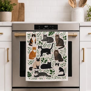May include: A white kitchen towel featuring a pattern of various cat breeds amidst green leaves and small pink hearts. The cats are depicted in different poses and colors, including black, orange, and tabby patterns. The towel is hanging on a gold-colored oven handle.