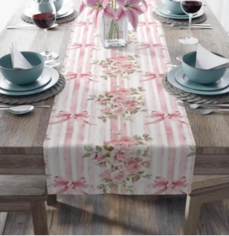 May include: A pink and white striped table runner with a floral pattern. The runner features pink roses and bows.