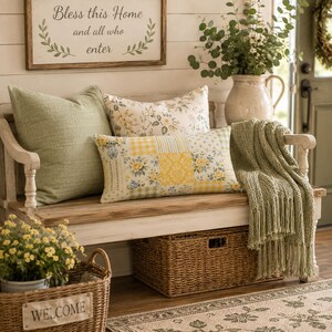 Pu&ograve; includere: Una panca in legno con base bianca presenta un cuscino verde salvia a tinta unita, un cuscino con stampa floreale e un cuscino rettangolare patchwork giallo e bianco. Un plaid in maglia verde salvia con frange pende di lato e un cesto &egrave; sotto.