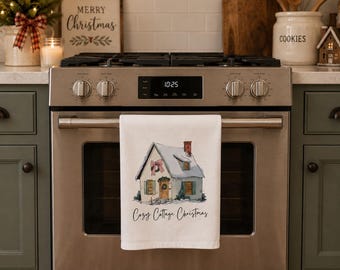 Puede incluir: Un paño de cocina blanco colgado en la puerta de un horno de acero inoxidable, con una ilustración en acuarela de una cabaña cubierta de nieve con un lazo rosa y el texto cursivo negro Cozy Cottage Christmas impreso bajo la casa.
