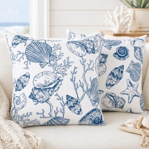May include: Two white throw pillows with a blue seashell and coral pattern. The pillows feature various seashell illustrations, including scallops, conchs, and starfish, creating a coastal theme. The pillows are placed on a white sofa.