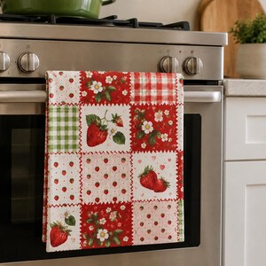 May include: A rectangular kitchen towel featuring a patchwork design of red strawberries, white strawberry blossoms, and green leaves on a white background, interspersed with red and white gingham and green and white checkered square patterns.