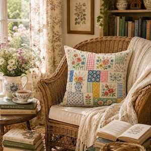 K&ouml;nnte beinhalten: Ein Patchwork-Kissen mit Blumenmustern liegt auf einem Korbstuhl, erg&auml;nzt durch eine cremefarbene Decke. Eine Blumenvase, B&uuml;cher und eine Teetasse verst&auml;rken die gem&uuml;tliche, Vintage-&Auml;sthetik. Das Kissen zeigt florale Designs in Rosa, Blau und Gelb.