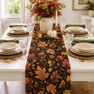 May include: A black table runner featuring a vibrant pattern of autumn leaves in shades of orange, red, yellow, and green. The runner is centred on a white dining table set with white plates, green napkins, gold cutlery, and clear glasses.